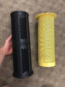 A yellow metal mesh filter beside the black 3D-printed honeycomb filter that replaced it, held in hand on a shop floor.
