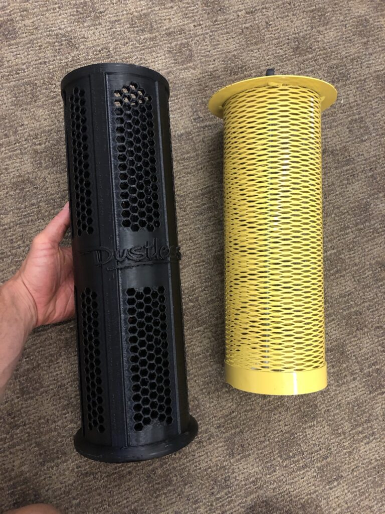 A yellow metal mesh filter beside the black 3D-printed honeycomb filter that replaced it, held in hand on a shop floor.