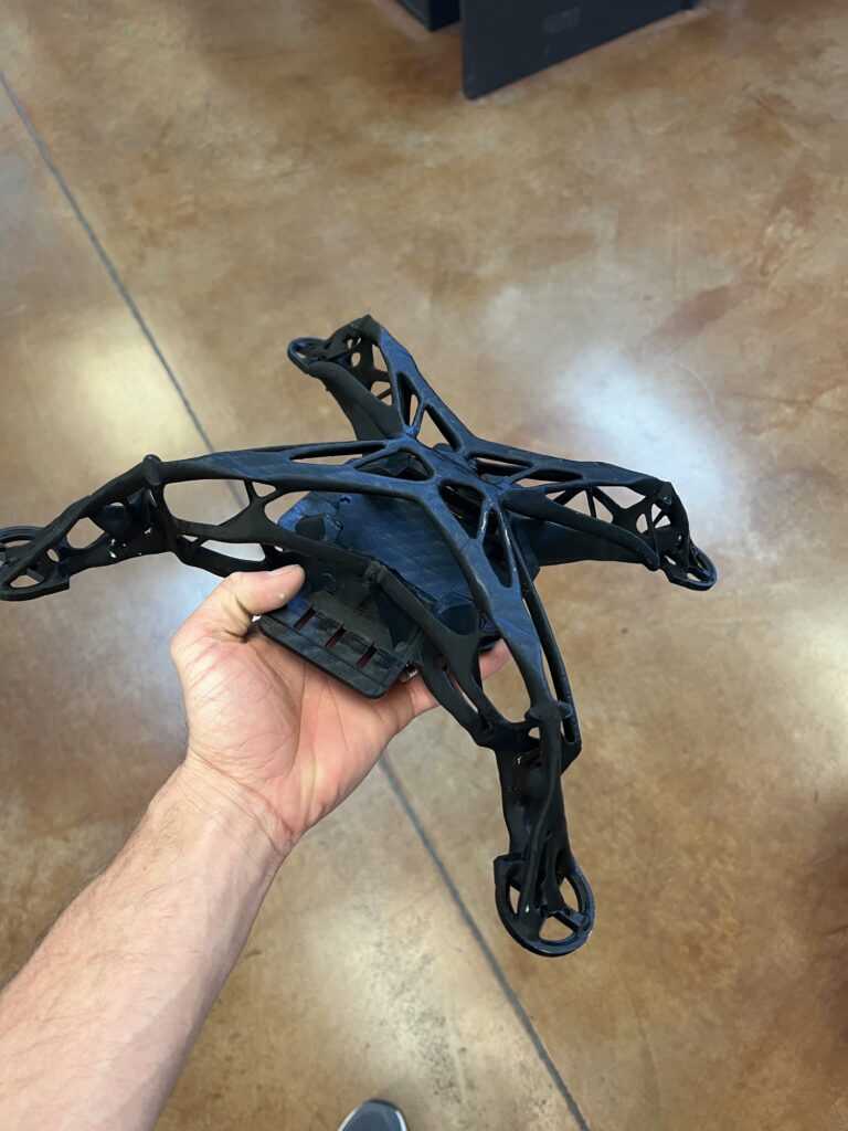 A 3D-printed drone frame with an organic lattice structure printed as one continuous part, held in hand on a shop floor.